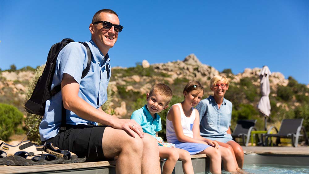 A man two children and a woman sitting at the edge of a pool with their feet in the water in the sun and smiling