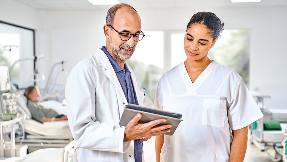 A nephrologist showing a dialysis nurse something on an iPad in a treatment room at a renal care center, with a patient in the background.
