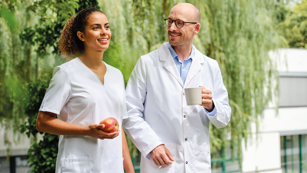 Two medical professionals, a dialysis nurse and a nephrologist, in white lab coats walking in front of dialysis center—one holding an apple, the other a white mug—with greenery and a building in the background.