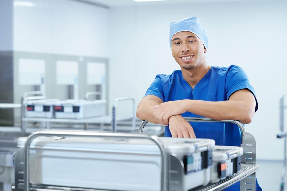 Healthcare professional in red surgical attire and cap standing in a modern sterile supply area, pushing a cart loaded with sealed sterile containers.