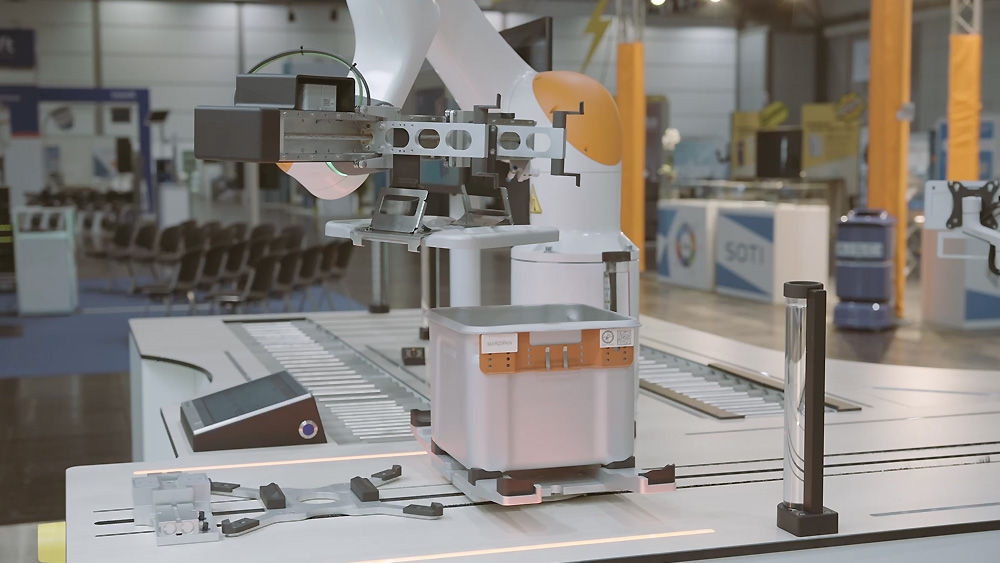 Industrial robotic arm operating on a sterile container in a modern automation setup, displayed at a trade fair with conveyor elements and precision tools on the workstation.
