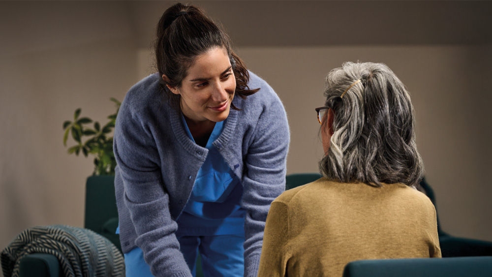 Two people are positioned in a seating area with armchairs; one person is leaning slightly forward while the other is seated, indicating an interaction in an indoor space.
