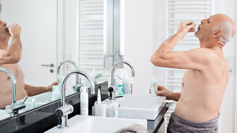 elderly man performing  antimicrobial mouth rinsing at home in front of bath mirror 