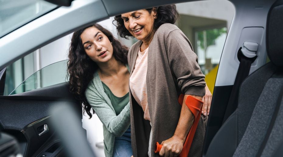 Young lady helps a middle-aged lady with crutches into the passenger seat of a car.