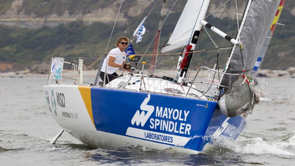 A man is sailing on a yacht through fast-moving waters, with waves splashing against the hull