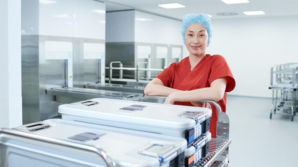 Healthcare professional in red surgical attire and cap standing in a modern sterile supply area, pushing a cart loaded with sealed sterile containers.