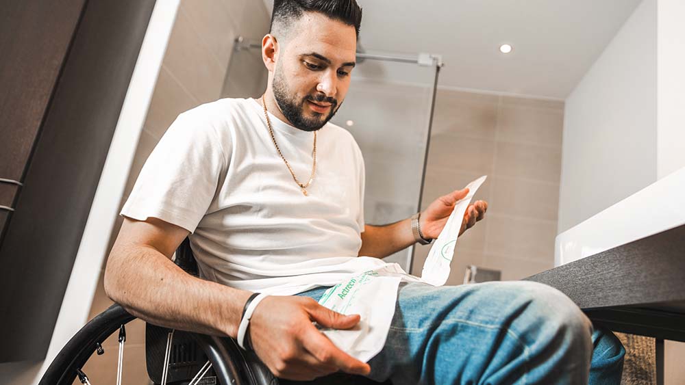 A man in a wheelchair, a bathroom in the background, holding a cathether in his hand