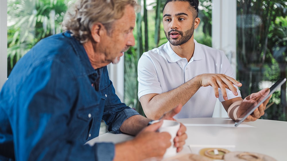 An old man with coffee and a young man with a tablet sit at a table and discuss.