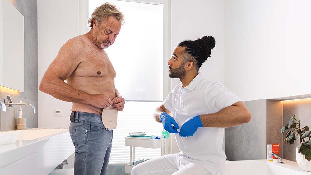 Man with bare upper body in private bathroom manipulates stoma bag on his stomach. Male nurse sits on the edge of the bathtub and gives instructions.