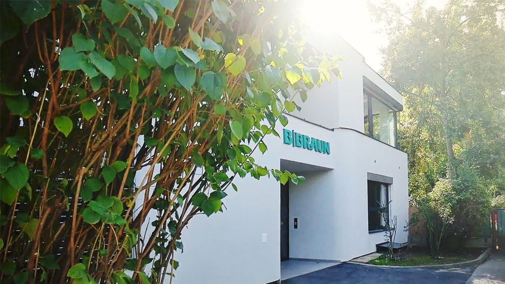 Modern white building with the green B. Braun logo on the facade, partially shaded by trees and greenery, with sunlight shining from the top right corner. The entrance and large windows are visible, and the setting appears to be a landscaped outdoor area.