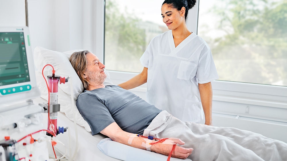 A nurse stands next to a patient undergoing dialysis, looking at him with care and compassion.