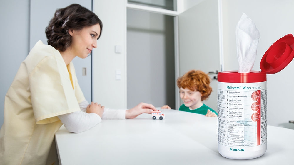 Female nurse standing at a table, opposite a small smiling boy. On the table is a small model ambulance being pushed by the woman, watched by the boy.  In the foreground a pack of Meliseptol Wipes.