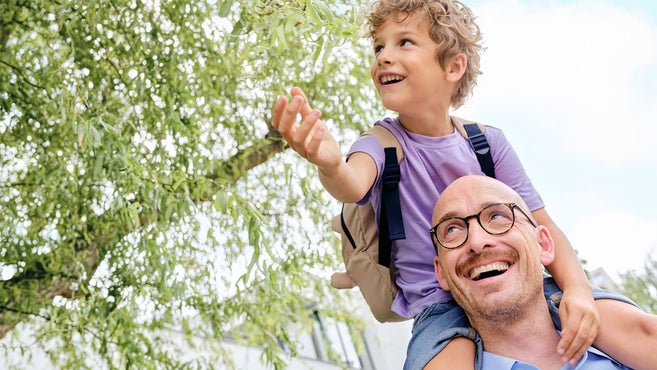 A child wearing a purple shirt and a backpack is sitting on an adult’s shoulders outdoors, reaching out with one hand to greenery. The adult is the childs father, he is a nephrologist.