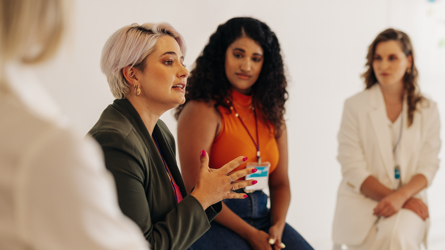 Four women are seated side by side in the Women's Mentoring Program. The goal is to increase the visibility of women in the organization and assist them in their personal growth. A second woman observes the first, a grey-haired woman in a green jacket, while she speaks and makes a hand signal.  