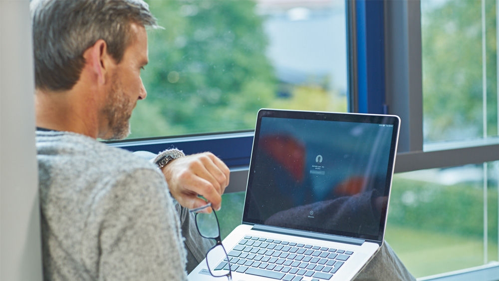 A seated person is holding a laptop on their lap. With their left hand, they are typing on the keyboard, while holding a pair of glasses in their right hand. On the laptop screen, a blurred symbol resembling headphones is visible, along with the label 'Tap to listen'. In the background, a green, natural landscape can be seen through a large window.