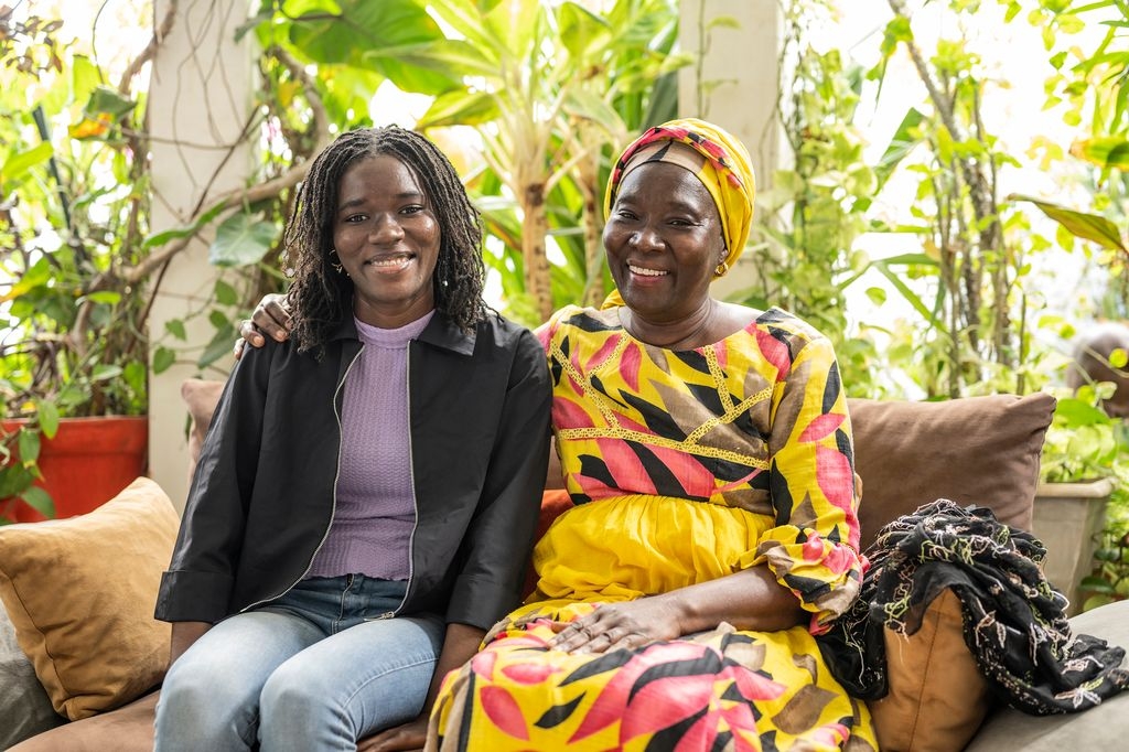 Silberia Diack and her mother are seated on a sofa in a well-lit room. They are holding each other in their arms and smiling facing forward.