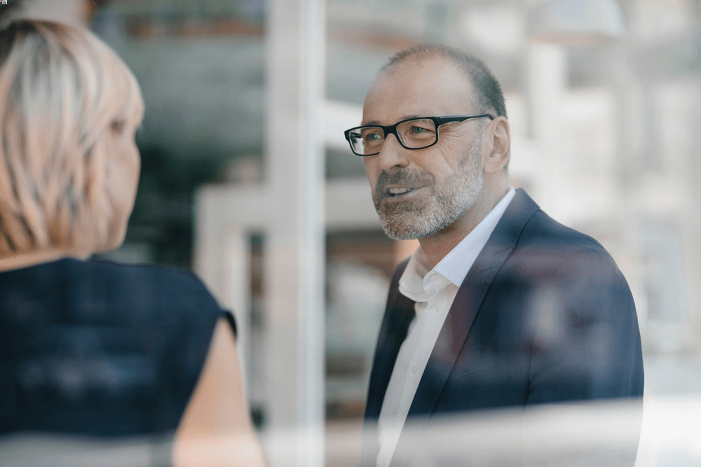 A man with glasses talks to a woman of whom you can only see the back of her head and her blond hair, the background is blurred.