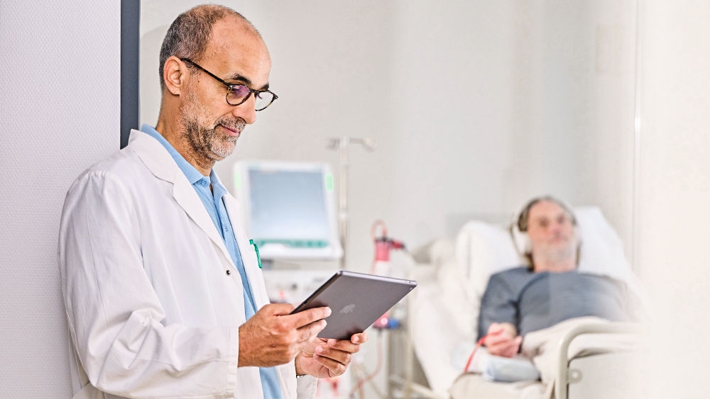 Healthcare professional using a tablet beside a patient receiving treatment in a hospital room, with medical monitoring equipment visible in the background.