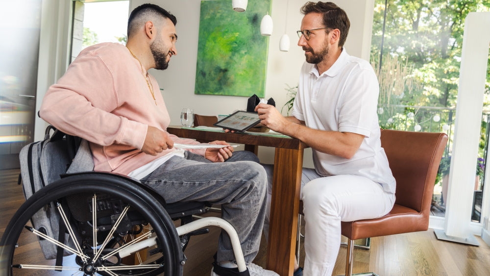 A man is sitting in a wheelchair, holding a catheter in his hands. In front of him, a hospital health specialist is giving instructions using a tablet