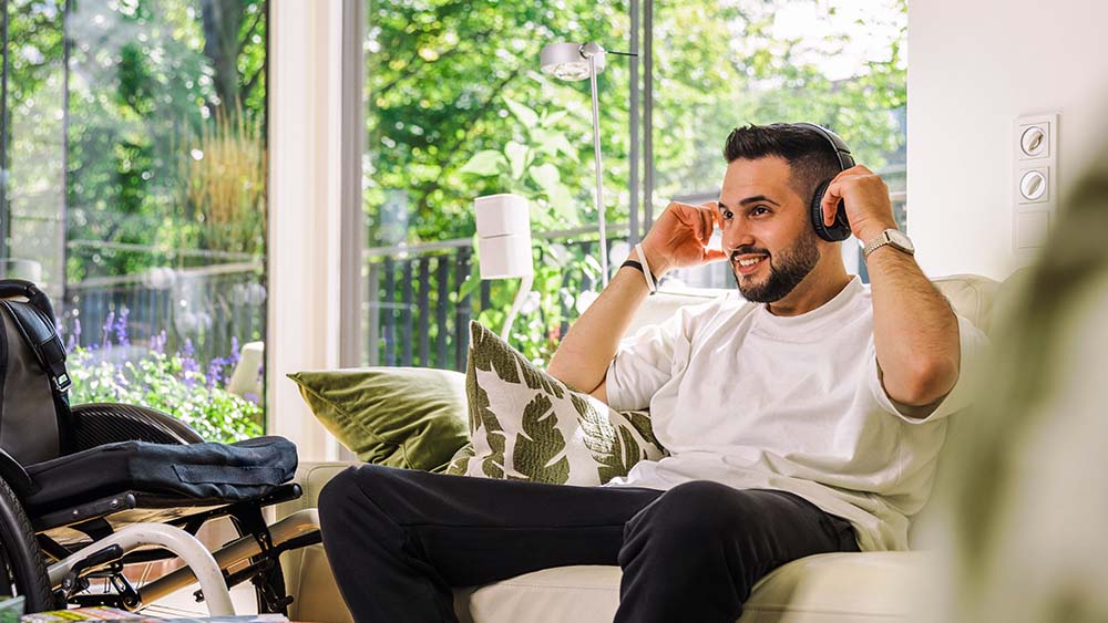 On the right a man is sitting on the sofa with headphones on, he is holding his hands on the headphones. To his left is a wheelchair