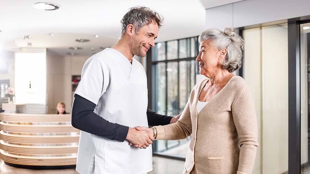 A doctor shakes hands with a patient, in the background is a reception area