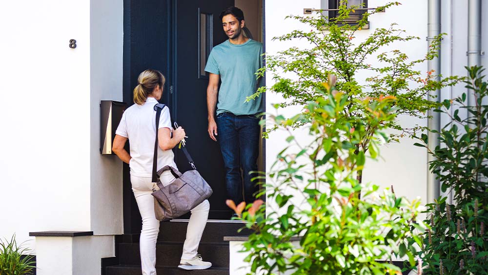 A nurse walks towards a staircase of a house door with a bag, a man stands in the open doorway to receive her