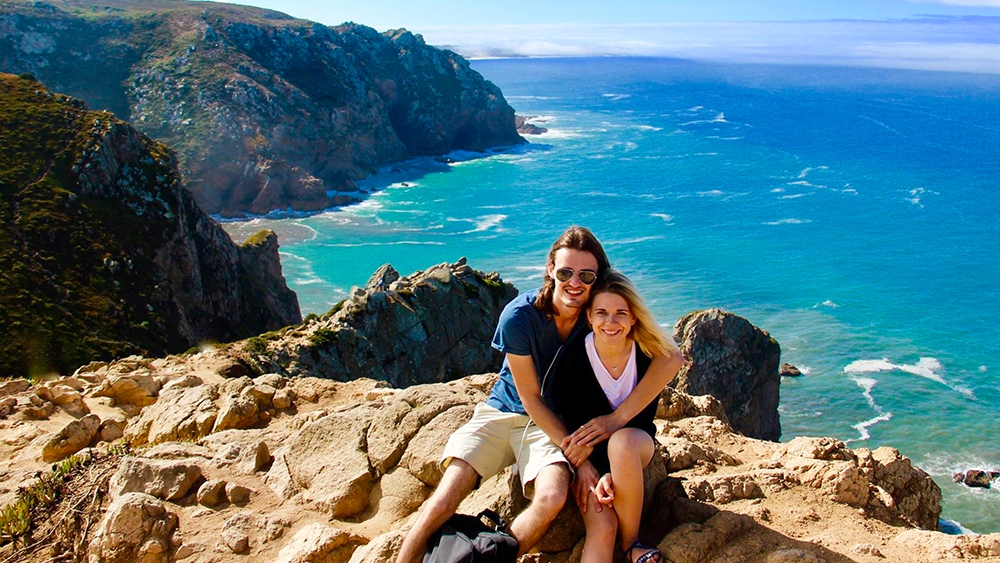 A man and a woman sit close together on a rock, smiling, with the sea in the background, their hair blowing in the wind