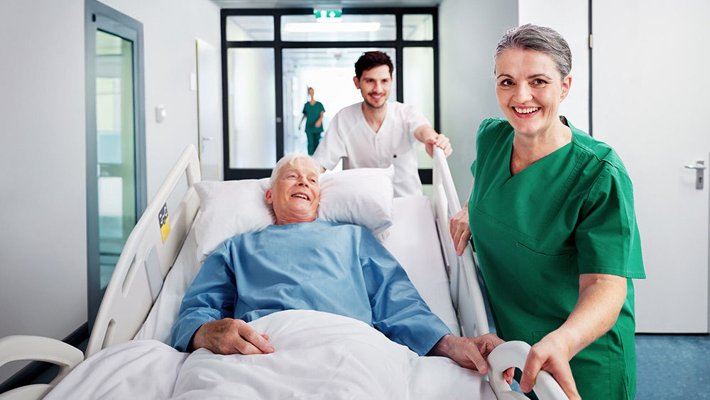 Patient lying in a hospital bed being transported through a corridor by health care staff, with another staff member walking alongside