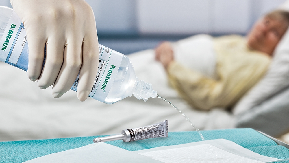 In the foreground, a hand wearing a medical glove pours Prontosan Irrigation Solution onto a pad lying on the table next to an Askina Calgitrol tube. In the background, a female patient lies in a hospital bed.