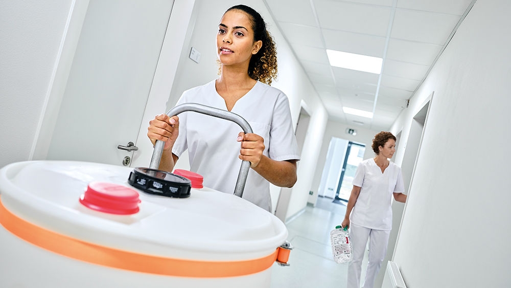 Two dialysis nurses in a clinical hallway. One pushes a ECOCart cartridge with dry concentrate powderon a trolley, while the other one walks in the background carrying a Sol-Can A Concentrate canister.