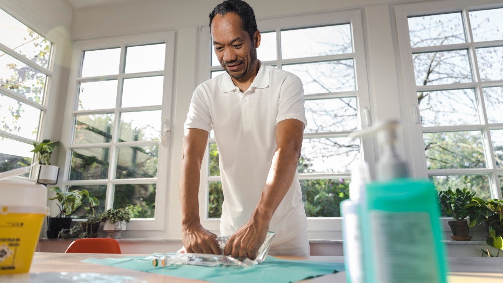 Male nurse prepares an infusion bag on a table in a brightly lit living room. Hand sanitizer can also be seen on the table.