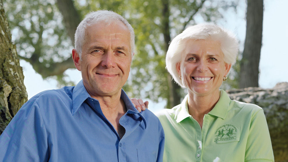 A middle-aged man and woman sit together in a forest, both smiling