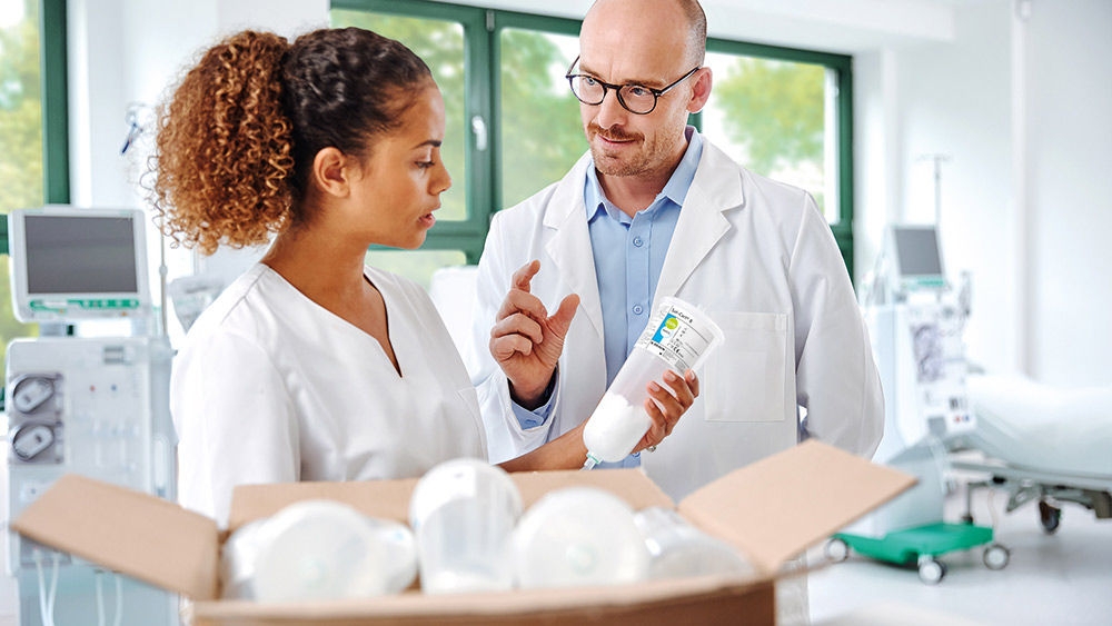 Two healthcare professionals, a dialysis nurse and a nephrologists, in a dialysis unit review a bicarbonate cartridge, with multiple cartridges prepared in an open box, highlighting resource‑conscious product selection in renal care.