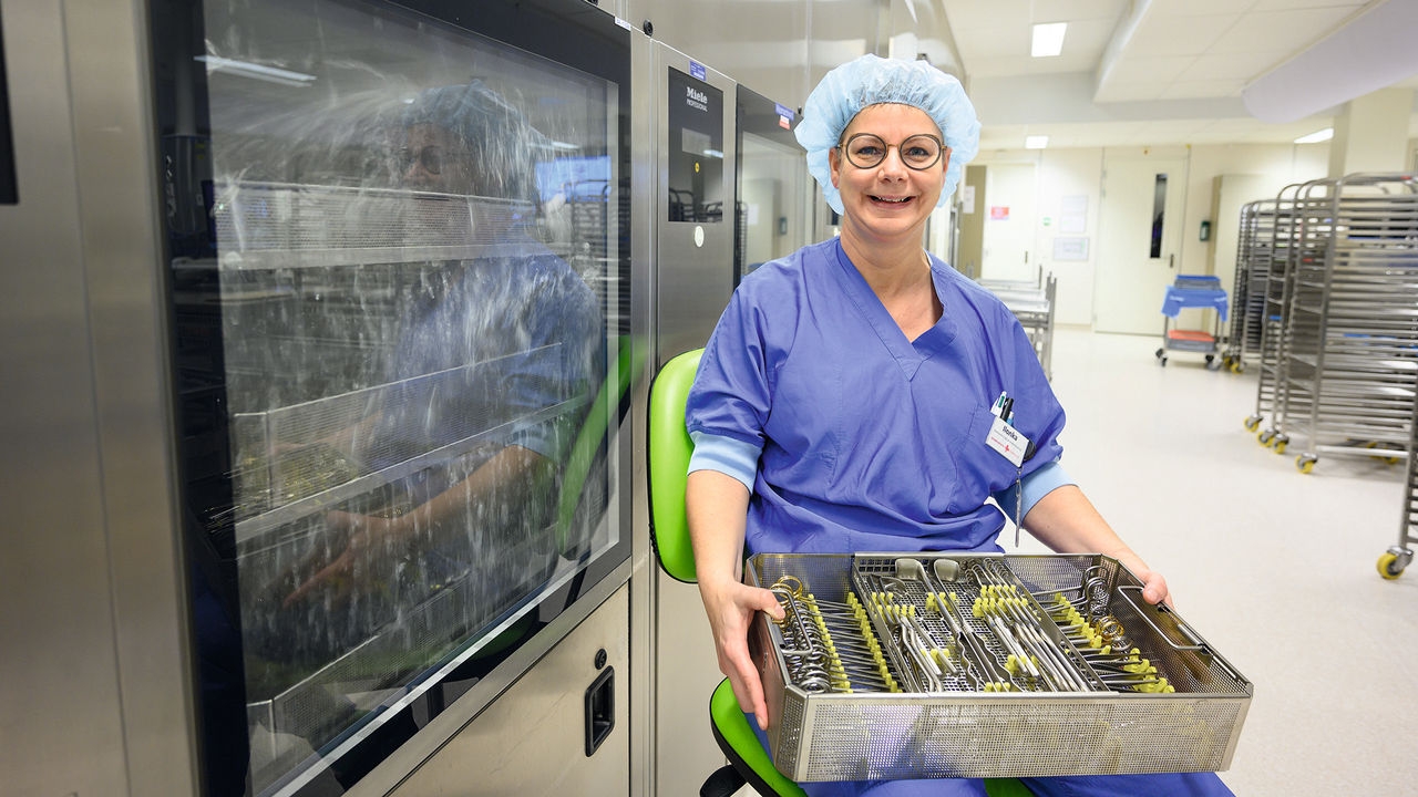 A smiling nurse sits on a chair with an instrument set, she is equipped with a hygiene cap