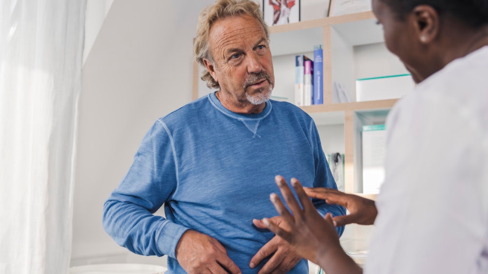 a middle-aged man asks the doctor a question while holding the right side of his stomach, a male doctor explains something to him