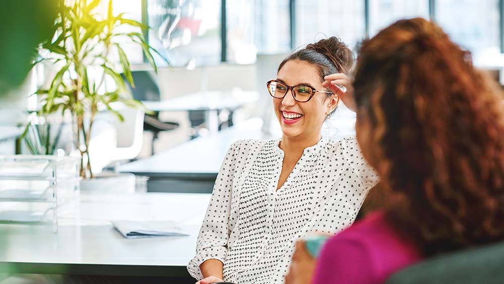 On the right sits a woman with the back of her head, on the left sits a woman in front of her who looks to the side and laughs. In the background is an office