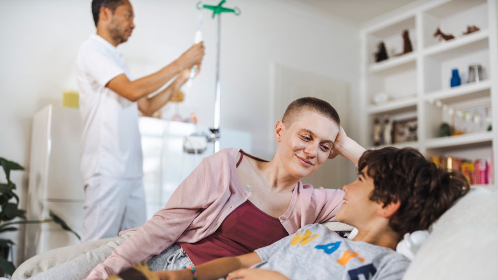 A woman with very short hair is lying next to a boy on a sofa, both with their upper bodies raised. They look at each other with a smile. In the background, a male nurse hangs an infusion bag on a rack.