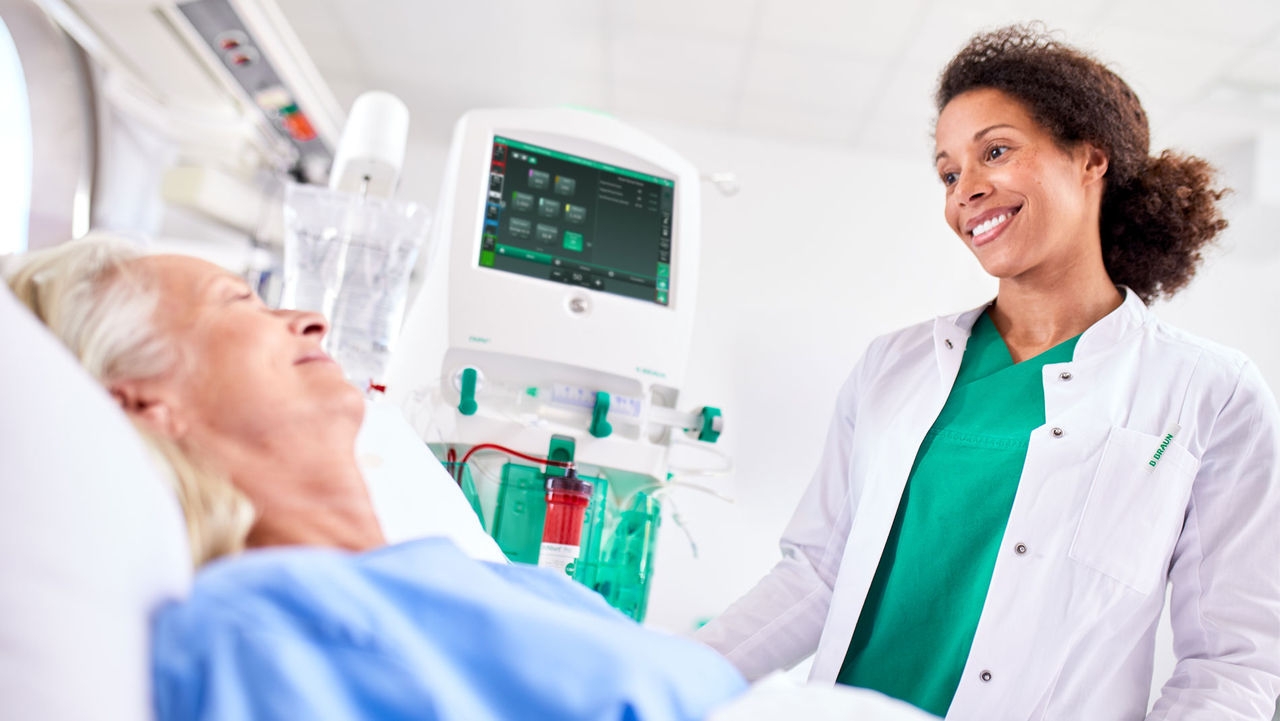 A  female physician stands in front of the patient's ICU bed. Both are smiling at each other. The OMNI machine is operating next to the bed