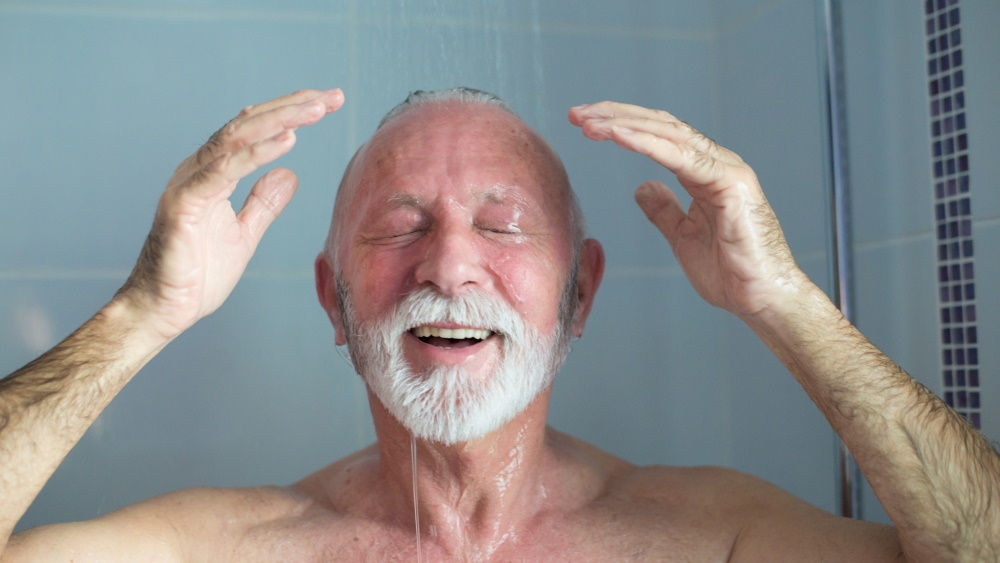 An elderly man is standing in the shower, smiling with the water running