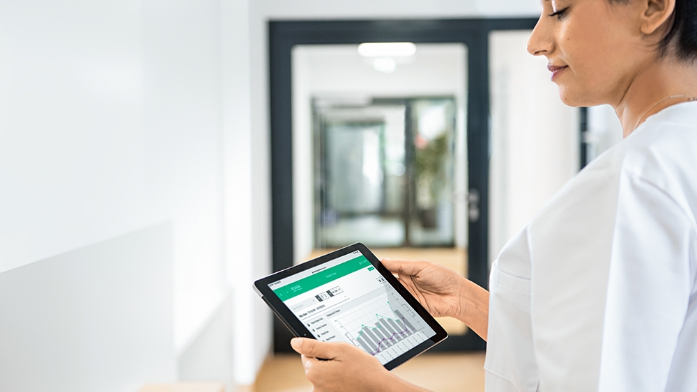 A famale medical worker holds a tablet with  NEXADIA® mobile companion open on it