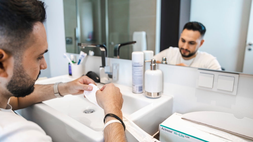 A man is sitting in front of a mirror in a private bathroom, holding a catheter in his hands.