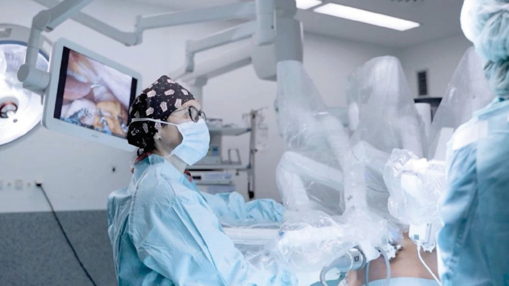Robotic-assisted surgery in progress, with a surgeon operating robotic arms while wearing surgical attire. In the background, an operating theatre light and a monitor displaying an internal view are visible.
