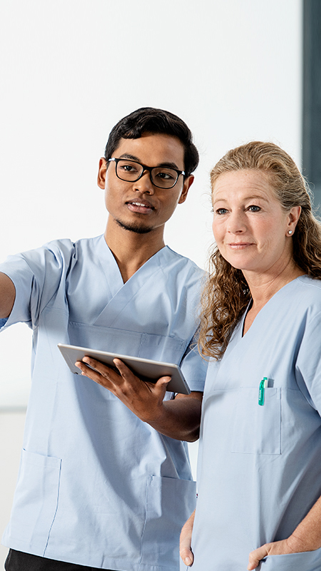 Two nurses, male and female, standing together. He has a tablet in his hand and is pointing forward with his other hand. Their eyes follow the direction.