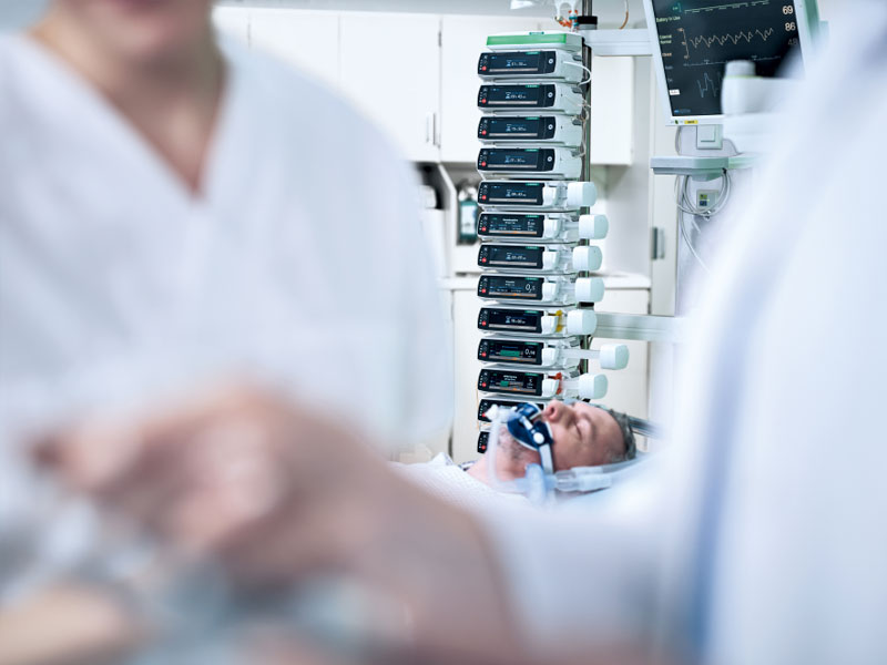 Two doctors are blurred in the foreground and in the background a patient lies in bed with a respirator mask and his eyes closed