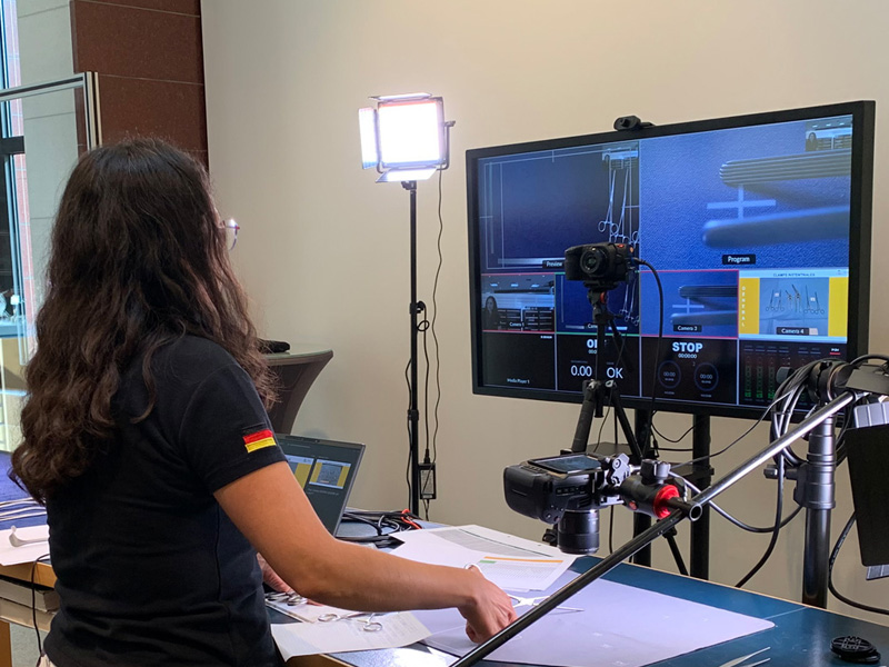 a woman stands in front of an office desk with two screens and a tripod with microphone, light and camera