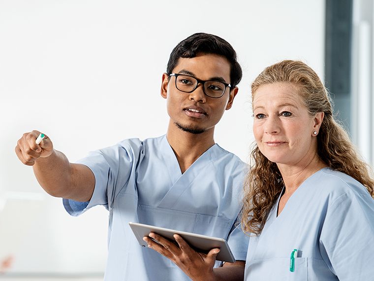 Two nurses, male and female, standing together. He has a tablet in his hand and is pointing forward with his other hand. Their eyes follow the direction.