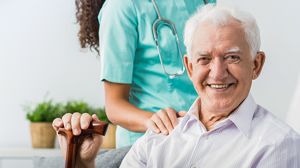 Elderly man sitting, sitting, laughing into the camera. Behind him nurse with hand on his shoulder.