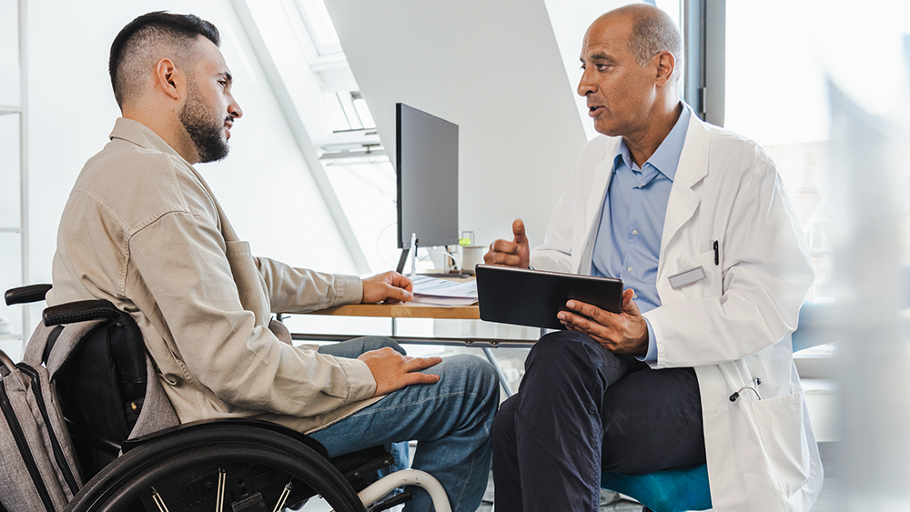 A man in a wheelchair sitting next to a doctor at his desk. the doctor explains something to him on an Tablet.