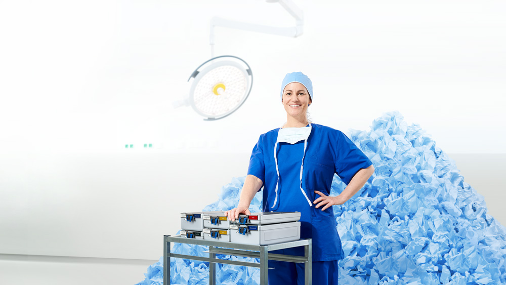 Healthcare professional standing beside sterile procedure packs in an operating room, with a large pile of blue disposable surgical materials in the background.