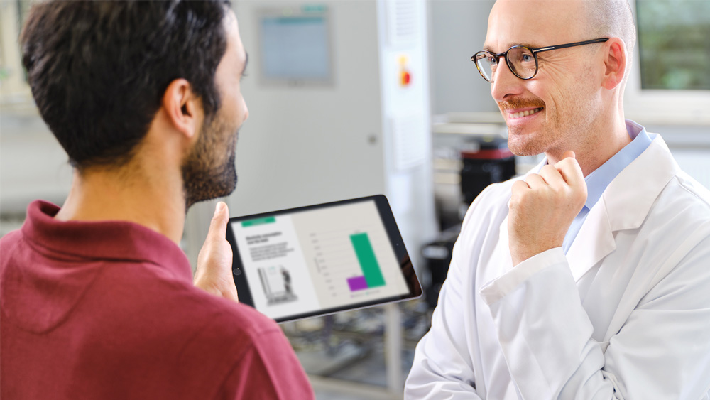 A dialysis technician and nephrologist in a professional setting infront of a water treatment system are discussing data displayed on a tablet. The tablet screen shows a bar chart with information about water and energy reduction in dialysis.